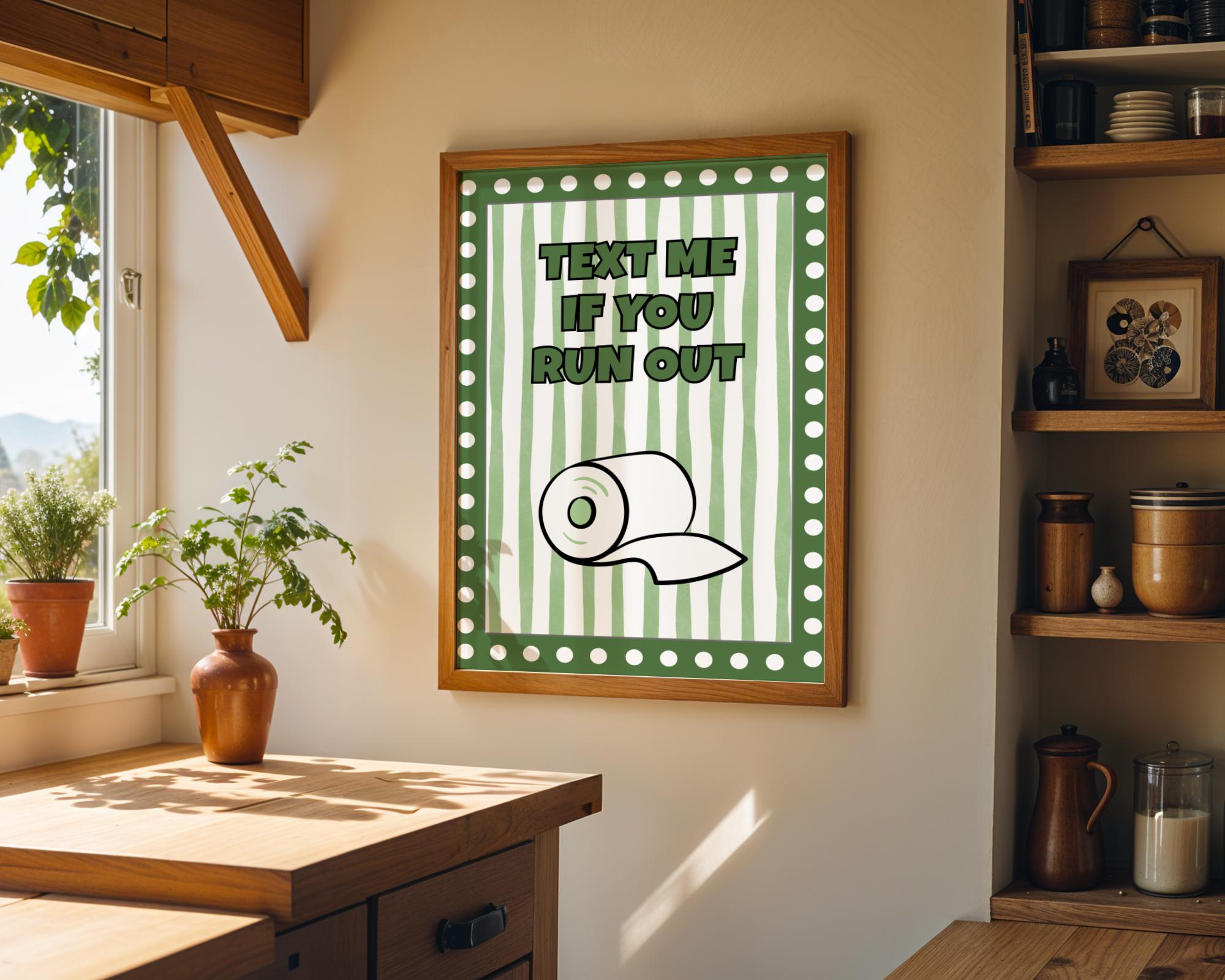 A framed wall art print hangs on a beige wall in a rustic kitchen setting. The print features a green and white striped background with the text 'TEXT ME IF YOU RUN OUT' and a simple line drawing of a toilet paper roll.