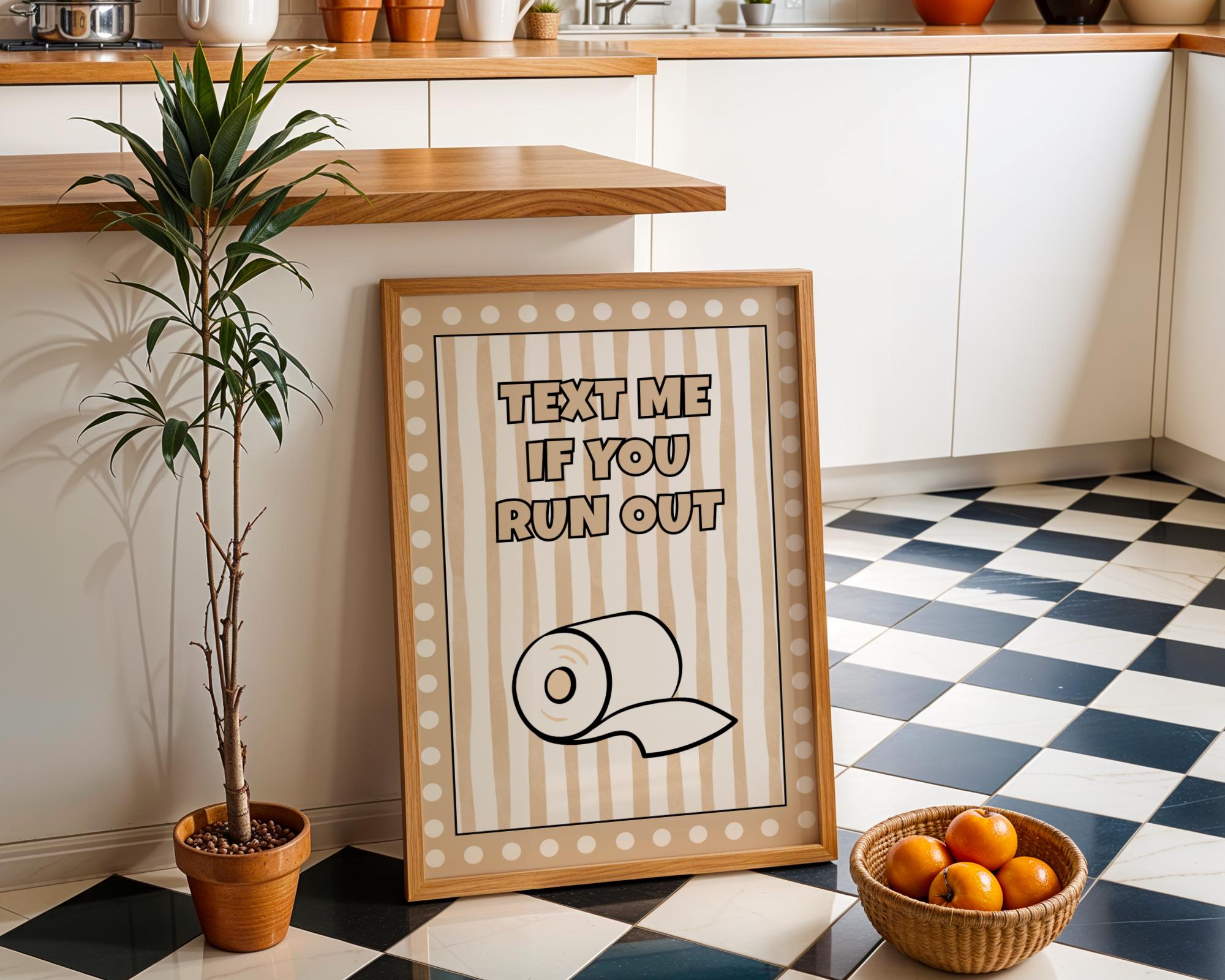 A framed novelty print with the text 'TEXT ME IF YOU RUN OUT' and a toilet paper roll illustration, leaning on a black-and-white checkered kitchen floor next to a potted plant and a basket of oranges.