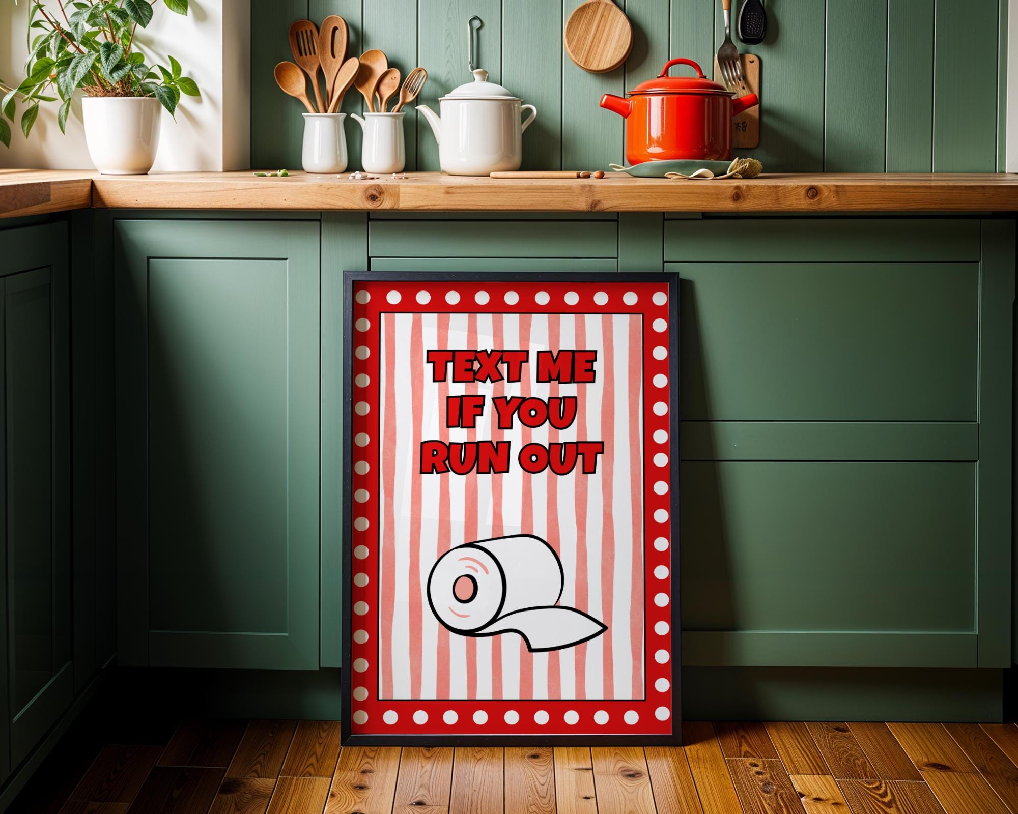 A framed novelty kitchen art print leaning against a green cabinet in a kitchen setting. The print features a red and white striped background with the text 'TEXT ME IF YOU RUN OUT' and a toilet paper roll illustration.