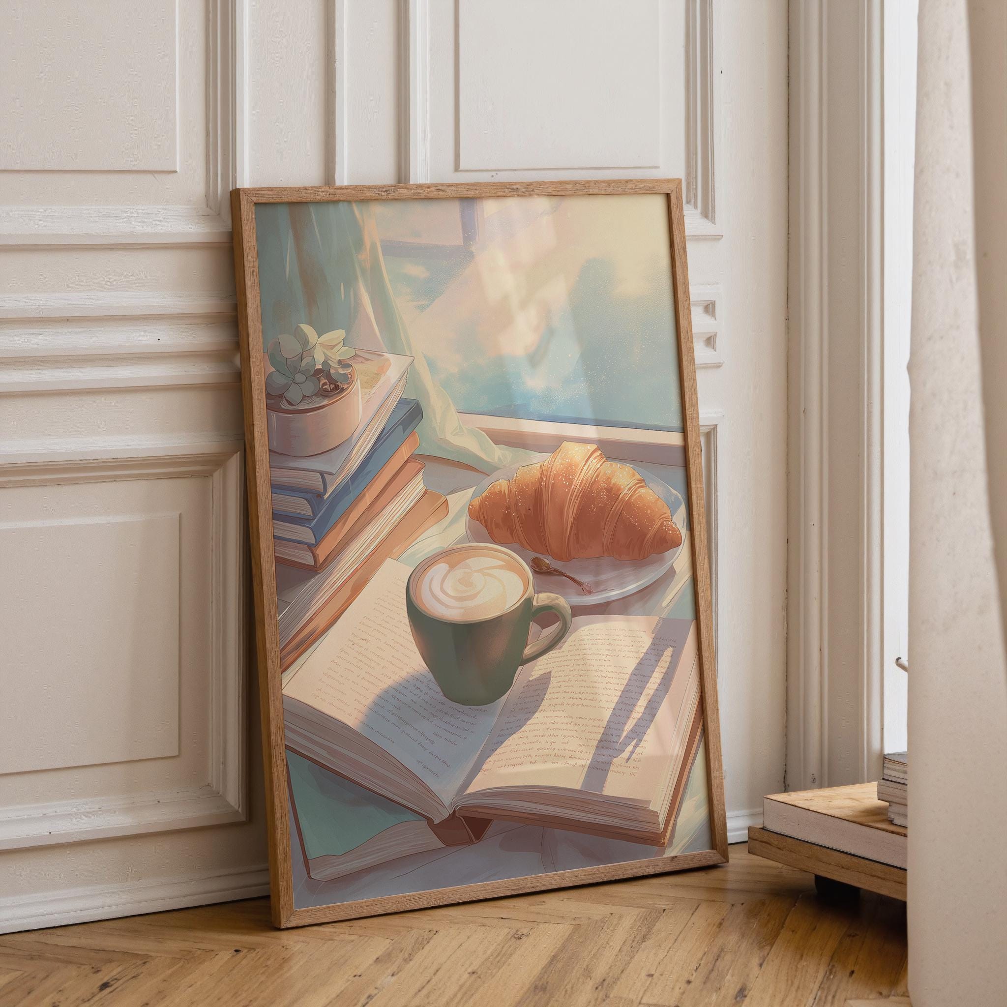 A framed art print leaning against a white paneled wall on a wooden floor. The print depicts a cozy still life scene with an open book, a green coffee cup with latte art, a croissant, and stacked books under soft light.