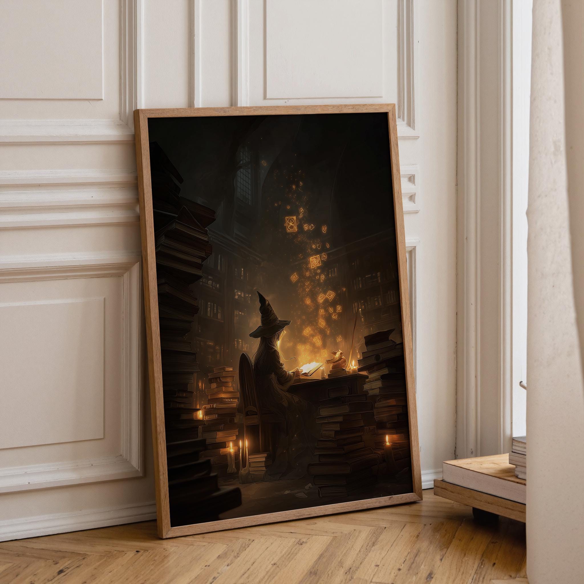 A framed vertical art print leaning against a white paneled wall on a light wood floor. The artwork depicts a dark, atmospheric scene with a witch-like figure in a pointed hat surrounded by glowing candles and stacks of books, with warm golden light and floating sparkles.