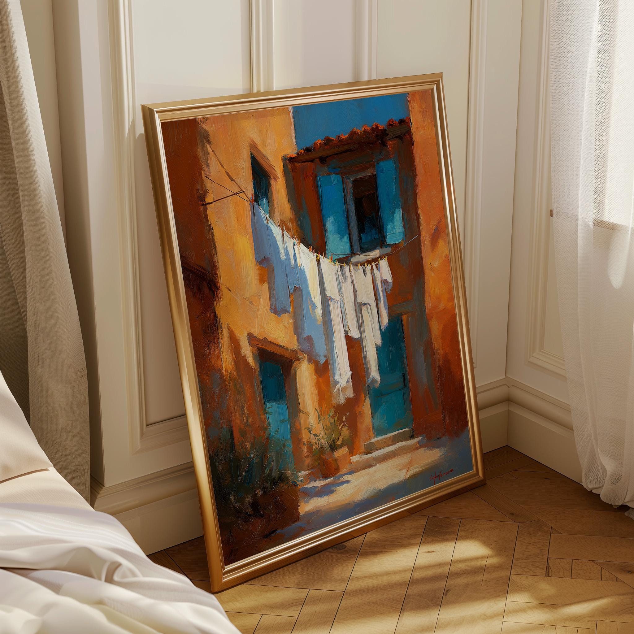 A framed painting of a Mediterranean-style street scene with warm orange and blue buildings, white laundry hanging on a line, and a gold frame, leaning against a white paneled wall on a wooden floor.